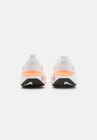 Weiße Sportschuhe mit orangen Akzenten, ausgestattet mit einem strukturierten Obermaterial, gepolstertem Kragen und einer ausgeprägten schwarzen Sohle mit Markenlogo.