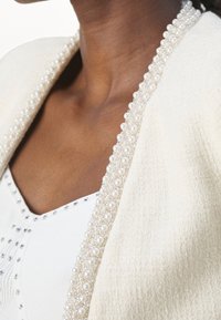 Gros plan d’une personne portant une veste blanche texturée avec un col orné de perles par-dessus un haut blanc avec de petits clous argentés.