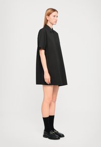 Black oversized shirt dress with short sleeves, button closure, and a relaxed fit. Worn with black socks and loafers. Smooth texture.