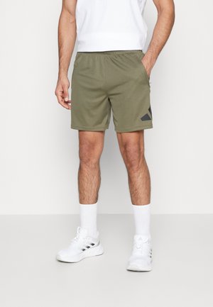 Homme portant un short de sport vert olive, des chaussettes blanches hautes et des baskets blanches avec des rayures noires, debout devant un fond clair uni.
