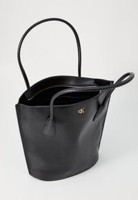 Sac fourre-tout en cuir noir avec poignées doubles, détail de logo et une fermeture éclair dorée. L'intérieur est doublé et spacieux avec une texture lisse.