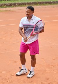 Tennis player in a gray and white polo with pink shorts, holding a pink racket, on a red clay court, wearing white socks and black shoes.