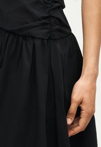 Κοντινό πλάνο ενός χεριού που ακουμπάει δίπλα σε μια πλισέ μαύρη φούστα με μαζεμένο ύφασμα στη μέση.