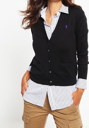 Femme portant un cardigan noir boutonné par-dessus une chemise rayée blanche et un pantalon cargo kaki, les mains dans les poches.