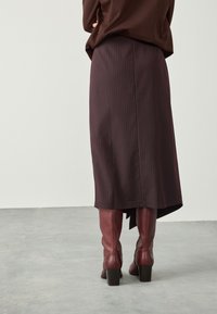 Jupe bordeaux à fines rayures avec un ourlet asymétrique, associée à des bottes à talons vernis couleur bordeaux. Texture de tissu douce ; design global épuré.