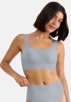 Vrouw met halflang donker haar, gekleed in een lichtgrijze naadloze beha en hoog uitgesneden ondergoed, tegen een effen achtergrond.