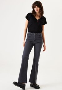 Zwarte v-hals t-shirt gecombineerd met hoog-geprijsde, uitlopende grijze jeans. Het model draagt grove zwarte schoenen, met een eenvoudige ketting als accent.