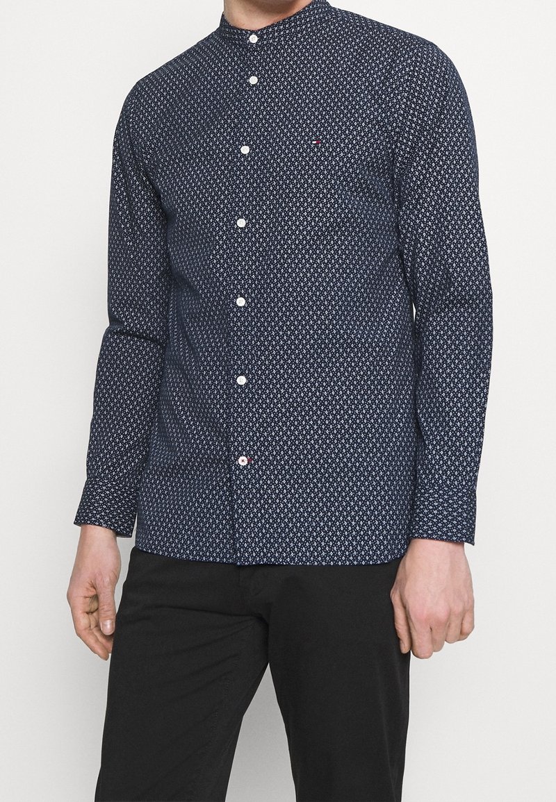 Camicia a maniche lunghe di colore blu scuro con motivo a diamante bianco, colletto alla mandarino e bottoni bianchi. Il tessuto appare liscio con una leggera lucentezza.