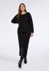 Top nero a maniche lunghe con scollatura decorata di borchie, abbinato a una gonna nera con spacco frontale e stivali neri con tacco al ginocchio.