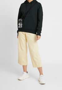 Sudadera de algodón negra con un bolsillo frontal, combinada con pantalones anchos de color amarillo claro. Zapatillas blancas y un bolso bandolera negro y gris con patrón.