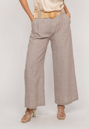 Femme portant un pantalon large en lin beige avec une ceinture tressée et des sandales à talons hauts dorées à brides, les mains dans les poches, debout devant un fond uni.