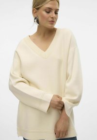 Pull crème en tissu doux, avec un décolleté en V profond, une coupe décontractée et un col côtelé, accompagné de manches longues légèrement fuselées.