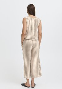 Femme portant un haut sans manches beige et blanc à carreaux avec des boutons dans le dos, et un pantalon large court assorti, chaussures noires glissantes.