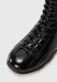 Černá syntetická kožená bota s kulatou špičkou, výrazným švem a šňůrkovým zapínáním. Má strukturovaný povrch a polstrovaný límec.