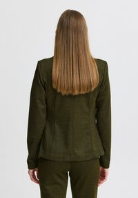 Dunkelgrüne maßgeschneiderte Jacke mit Fischgrätenmuster, strukturierten Schultern, schlanker Passform und langen Ärmeln, kombiniert mit passenden Hosen.