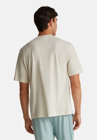 Muž v jednobarevném světlém béžovém tričku s krátkým rukávem a světlých modrých kalhotách, zachycený zezadu na bílém pozadí.