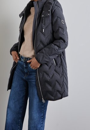 Persoon draagt een zwarte doorgestikte winterjas over een beige trui en blauwe spijkerbroek, staat met handen in de zakken tegen een grijze achtergrond.