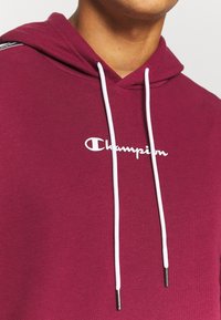Primo piano di una persona che indossa una felpa Champion maroon con cordini bianchi e logo sul petto.