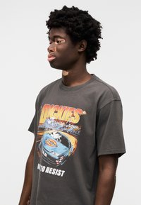 Γκρι βαμβακερό μπλουζάκι με τυπωμένο γραφικό μπλέ αγωνιστικό αυτοκίνητο και έντονο κείμενο "DICKIES" στο μπροστινό μέρος. Σχεδιασμένο με κοντά μανίκια και λαιμόκοψη τύπου crew.