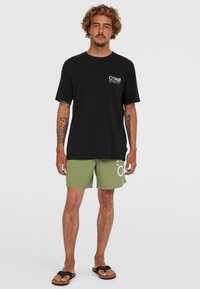 T-shirt en coton noir avec un logo blanc sur la poitrine, associé à un short vert olive avec un graphique blanc. Porté avec des sandales noires.