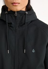 Black hooded jacket with a front zipper, drawstrings, and silver hardware. Features a subtle logo on the left chest. Smooth fabric texture.