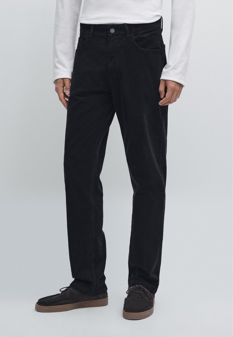 Svarta manchesterbyxor med rak passform, med framfickor och en enkel knäppning. I kombination med mörka mockaskor.