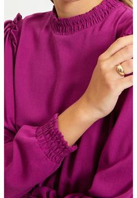 Haut à manches longues violet avec un col et des poignets froncés, en tissu lisse. Le design comprend des accents subtils de volants près des épaules.