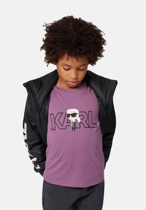 Paarse katoenen T-shirt met een cartoonafbeelding en vette tekst "KARL." Het kind draagt een zwarte zip-up jack met witte accenten.