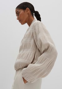 Hellbeige strukturierte Jacke mit horizontalen Falten, fallenden Ärmeln und lockerer Passform, kombiniert mit einem glatten, eng anliegenden cremefarbenen Oberteil.