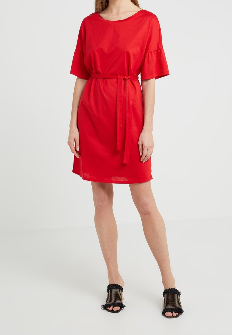 Vestido de algodón rojo con mangas cortas con volantes, un lazo en la cintura y un corte holgado, combinado con zapatos negros con tiras que tienen bordes deshilachados.