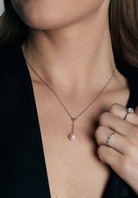 Srebrny naszyjnik łańcuszkowy z pojedynczą perłą w kropli, akcentowany małymi srebrnymi koralikami, noszony na tle czarnego materiału.