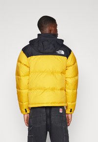 Puffertjakke med en gul krop og en sort øverste sektion, hætte og The North Face-logo. Struktureret, quiltet design med elastiske manchetter.