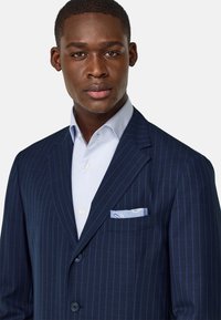 Veste de costume bleu marine à rayures avec une chemise habillée bleu clair. Elle présente des revers en pointe, deux boutons et un mouchoir de poche à motif cachemire.