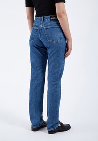 Vaqueros de mezclilla azul con corte recto, que cuentan con dos bolsillos traseros y una etiqueta de marca negra en la cinturilla. Textura suave y diseño clásico.