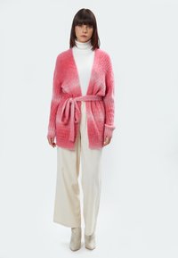 Roze gradient cardigan met een striktaille, geribbeld textuur, open voorkant en zachte stof, gecombineerd met crèmekleurige broek met wijde pijpen en glanzende laarzen.