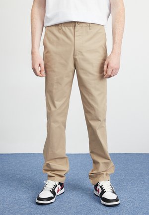 Mann trägt beige gerade geschnittene Hose und schwarze, weiße und rote Turnschuhe und steht auf blauem Teppich.