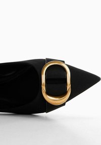 Chaussure à talon haut en satin noir avec un bout pointu et une grande boucle ovale dorée sur la bride avant. Texture lisse, design épuré.