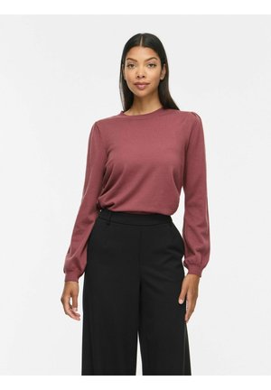 Kvinde med langt mørkt hår iført en mauve langærmet top og sorte, højttaljede, vidde bukser stående mod en hvid baggrund.