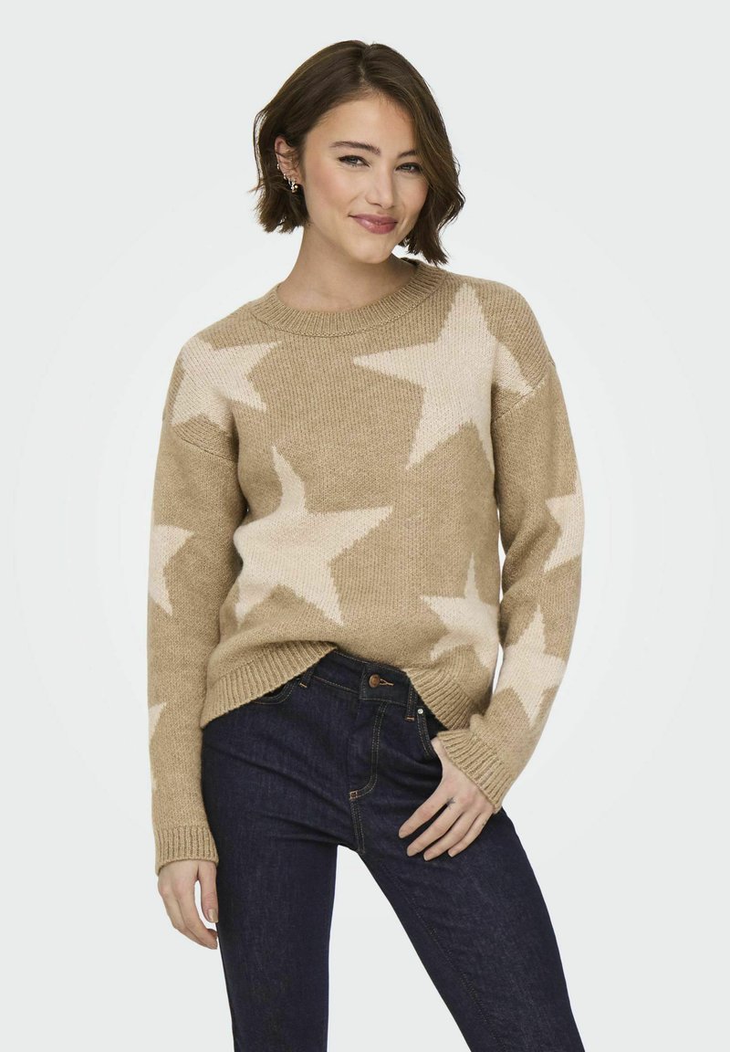 Beige gebreide trui met groot crèmekleurig sterrenpatroon, voorzien van een ronde halslijn, geribde manchetten en zijsplitten. G gedragen met donkere jeans.