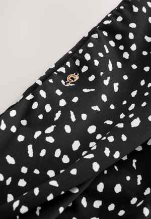 Zwarte stof met een wit gestippeld patroon en een klein, goudkleurig embleem. De textuur lijkt glad en licht reflecterend.