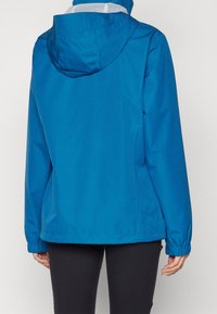 Veste imperméable bleue avec capuche, en tissu texturé, poignets élastiqués et silhouette ample. Vue de dos montrant des lignes épurées.