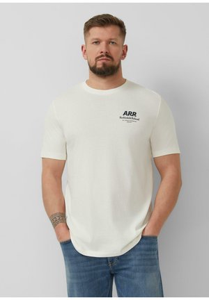 Brodaty mężczyzna z krótkimi włosami, ubrany w biały T-shirt z nadrukiem A.R.R. i niebieskie jeansy, stoi z rękami w kieszeniach na jednolitym tle.