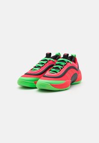 Des chaussures de sport rouge vif et vert présentent une tige en mesh texturé, des accents noirs bien visibles, un rembourrage épais et des lacets verts.