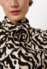 Zwarte en crèmekleurige blouse met een strikkraag, voorzien van golvende, abstracte patronen en een textiel met een structuur. Close-up van de halslijn.