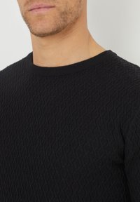 Pull en maille noir avec un motif en diamant texturé et un col rond. Le tissu semble doux avec une coupe près du corps.