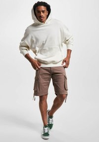 Kremfarget hettegenser med kengurulomme, kombinert med lysebrune cargo shorts med lommer på sidene. Grønn og hvit sneakers fullfører antrekket.