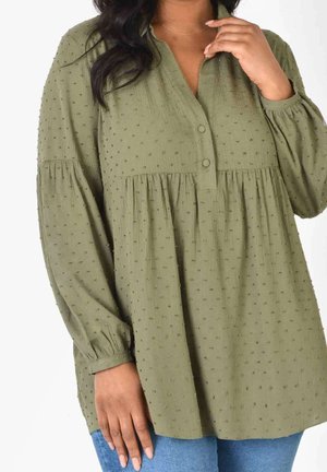 Frau trägt olivgrüne strukturierte Langarmbluse mit Knöpfen und blaue Jeans, Hand nahe am Kragen und seitlich herab.