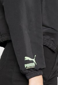Juoda striukė su elastinėmis rankogaliais, ant rankovės esantis žalias Puma logotipas. Medžiaga atrodo lygi su matinio paviršiaus apdaila.