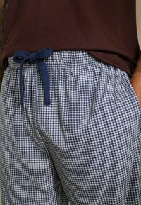 Kalhoty vzoru houndstooth v modro-bílé kombinaci s námořnickou modrou šňůrkou uvázanou do mašle, nošené s tmavě hnědým tričkem zastrčeným lehce dovnitř.