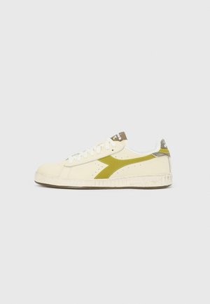 Sneaker basse crème avec lacets blancs, bande latérale vert olive, semelle blanche texturée et détail marron sur la languette et la languette arrière.